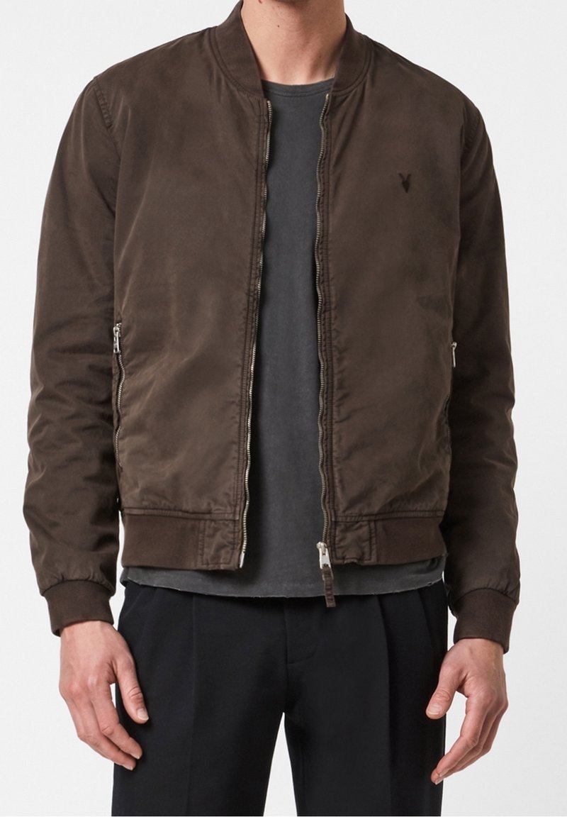 Homme portant une veste bomber marron à fermeture éclair sur un t-shirt gris foncé avec un pantalon noir, sur un fond uni.