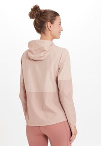 Kevyt, vaaleanpunainen tuulenpitävä takki hupulla, jossa on väriestetiikka ja elastiset hihansuut. Sileä rakenne ja rento istuvuus.