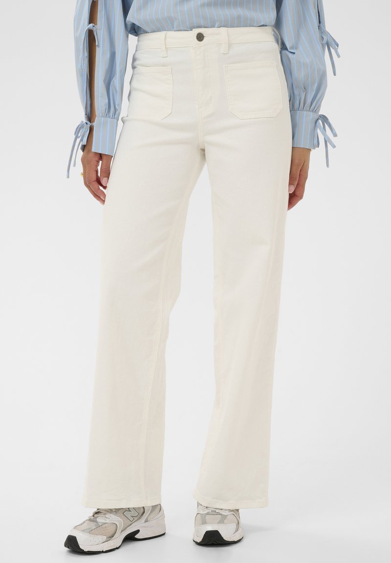 Pantalon blanc taille haute à jambes larges avec poches avant, porté avec des chaussures de sport grises et blanches, porté par une personne portant une chemise à rayures bleues.