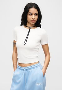 Λευκό ριμπ crop top με στρογγυλή λαιμόκοψη και λεπτομέρειες με μαύρο κορδόνι. Συνδυασμένο με ανοιχτό μπλε φόρμα, με λογότυπο στην τσέπη.