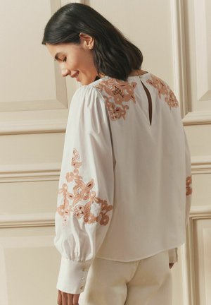 White blouse with long puffed sleeves, featuring coral floral embroidery on sleeves and back, and a small back keyhole detail.