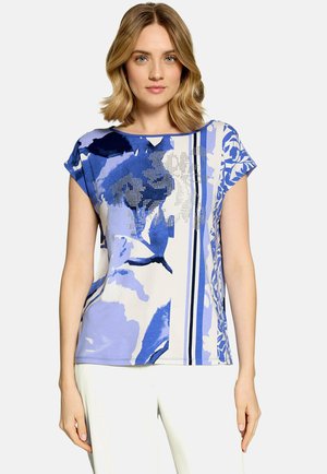 Femme portant un haut à manches courtes à motif floral bleu et blanc avec un pantalon blanc, debout devant un fond uni.