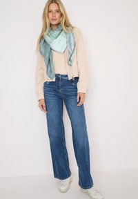 Chaqueta bomber beige con mangas elásticas, camiseta clara, jeans acampanados azules y una bufanda de patrón verde azulado. Zapatillas blancas completan el atuendo.