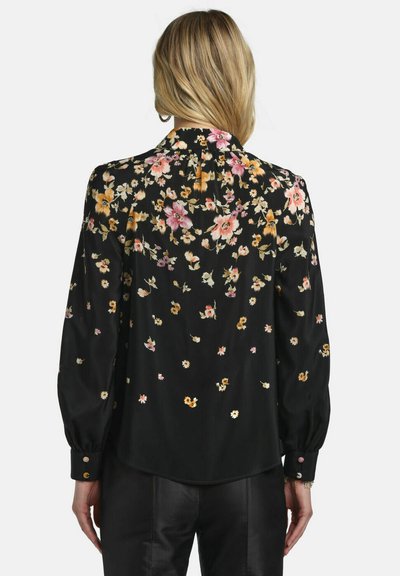 Femme portant un chemisier noir à manches longues avec un imprimé floral rose, blanc et jaune vue de dos, assorti à un pantalon noir.