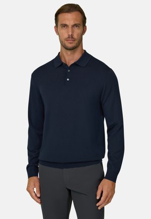 Homme portant un polo à manches longues bleu marine et un pantalon foncé, debout devant un fond uni et clair.