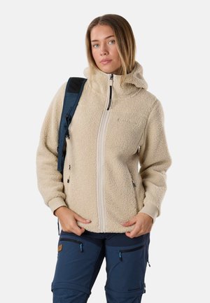 Kvinde iført beige fleecejakke med hætte og blå cargobukser, bærende på en navy rygsæk over den ene skulder.