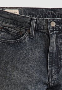 Gros plan sur un jean en denim foncé montrant la ceinture, la boucle de ceinture, la poche avant et le bouton avec une étiquette partiellement visible à l'intérieur de la ceinture.
