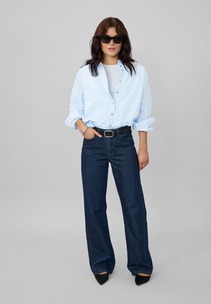 Femeie purtând o cămașă albastru deschis supradimensionată cu nasturi, blugi largi închiși la culoare, curea neagră, pantofi negri cu vârf ascuțit și ochelari de soare negri, stând cu o mână în buzunar.