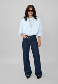 Light blue button-up shirt, dark denim wide-leg jeans, black belt, and pointed black shoes. Model wears dark sunglasses and stands confidently.