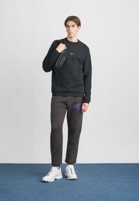 Un joven está de pie con una sudadera negra de Reebok, pantalones de chándal gris oscuro con un logo morado, zapatillas blancas y sosteniendo un bolso negro de hombro.