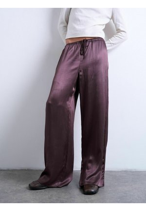 Løse, skinnende lilla satin bukser med vid ben, elastisk talje og snøre, båret med mørkebrune sko og en hvid cropped top.