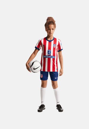 Kind in rot-weiß gestreiftem Fußballtrikot und blauen Shorts hält einen schwarz-weißen Nike-Fußball vor einfarbigem Hintergrund.