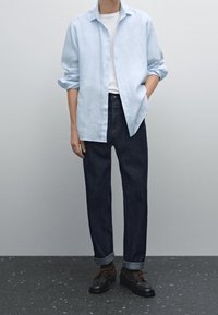 Camicia di lino azzurro chiaro a maniche lunghe sopra una maglietta bianca, abbinata a jeans in denim scuro e scarpe nere con lacci marroni.