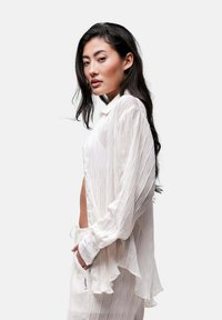 Camicia leggera bianca a maniche lunghe con un sottile motivo a righe verticali, caratterizzata da una chiusura con bottoni frontale e una vestibilità rilassata con orlo arricciato.