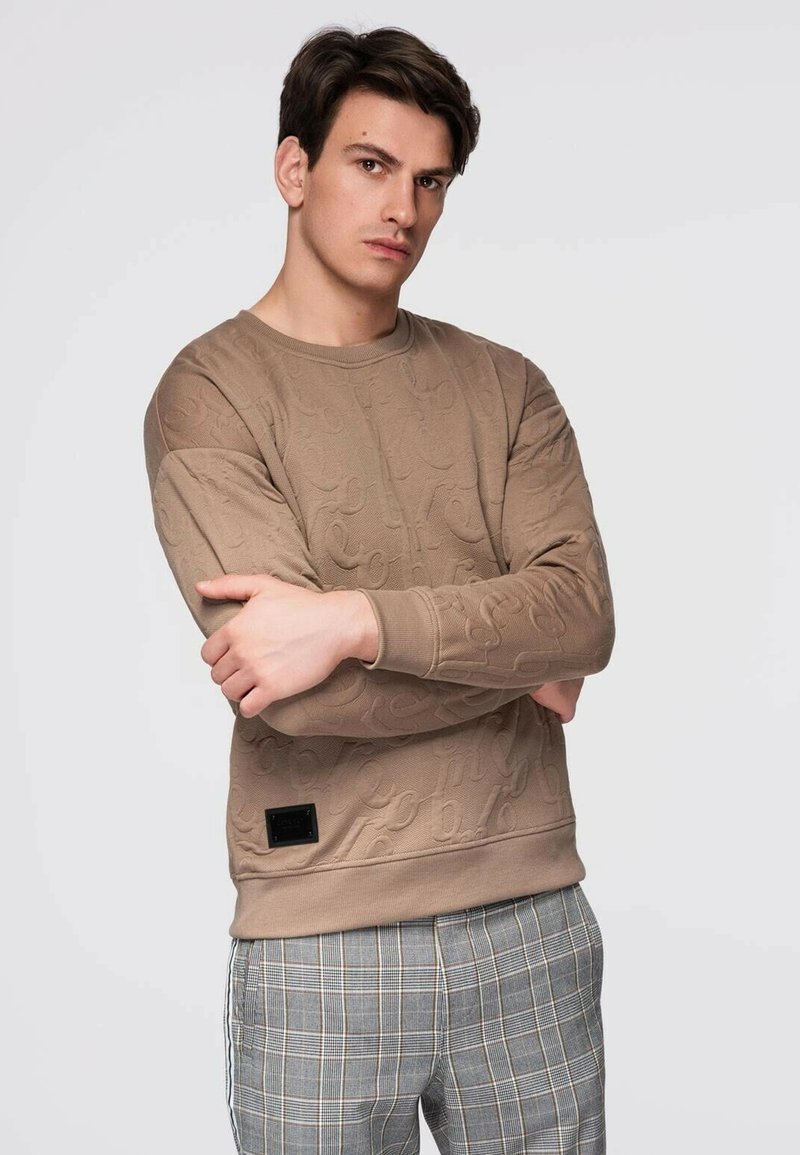 Beige collegepaita, jossa on teksturoitu kuvio, pyöreä pääntie, resorit hihansuissa ja musta etikettidetalji helmassa, yhdistetty harmaihin ruutukuosisiin housuihin.