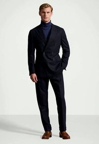 Άνδρας που φοράει σκούρο διπλό σακάκι με ναυτικό ζιβάγκο και καφέ δερμάτινα παπούτσια, στέκεται με το ένα χέρι στην τσέπη σε ουδέτερο φόντο.