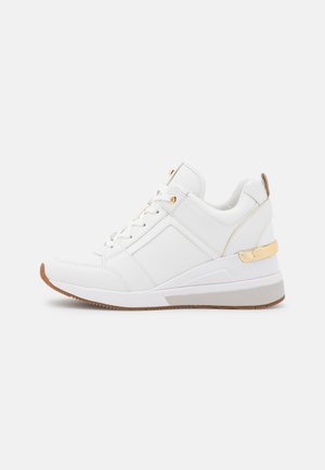 Biały niski sneaker z złotymi przelotkami i akcentem na pięcie, z przodu sznurowany, z beżową gumową podeszwą i lekkim klinowym obcasem.