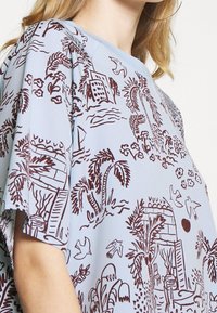 Ljusblå topp med brun tropisktryck, inklusive palmer, byggnader och fåglar. Avslappnad design med l ösa ärmar.