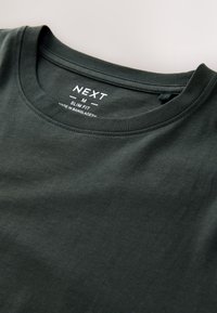 Temno zelena ozka majica iz bombaža, z okrolim izrezom in etiketo z informacijami o znamki, prišito ob ovratniku.