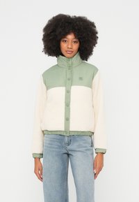 Volüümikate lokkis juustega naine, kes kannab kinnitatud rohelise-valge fliisjopet ja helesiniseid teksapükse, seisab tavalise valge tausta ees.