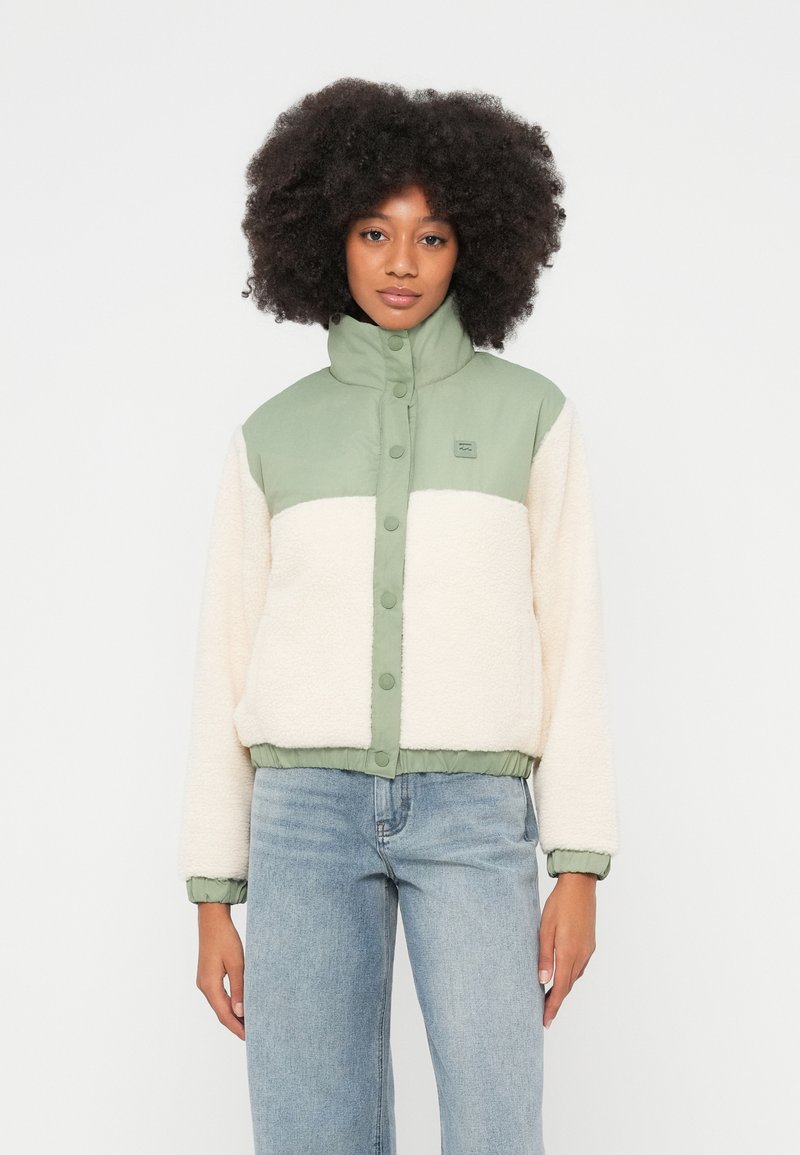 Volüümikate lokkis juustega naine, kes kannab kinnitatud rohelise-valge fliisjopet ja helesiniseid teksapükse, seisab tavalise valge tausta ees.