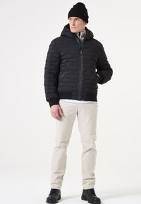 Schwarze Puffjacke mit Reißverschluss, Kapuze und gerippten Manschetten. Getragen über einem grauen Pullover und kombiniert mit hellen Hosen und schwarzen Stiefeln.