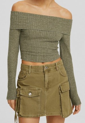 Femme portant un crop top côtelé olive vert à épaules dénudées avec manches longues et une mini-jupe cargo beige avec des poches à boutons.