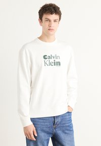 Biela mikina vyrobená z jemnej tkaniny, s výrazným zeleným logom "Calvin Klein" v tučnom písmene. S rebrovanými manžetami a lemom, vo voľnom strihu, v kombinácii s modrými džínsami.