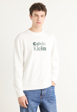 Biela mikina vyrobená z jemnej tkaniny, s výrazným zeleným logom "Calvin Klein" v tučnom písmene. S rebrovanými manžetami a lemom, vo voľnom strihu, v kombinácii s modrými džínsami.