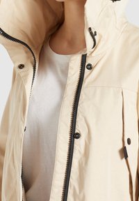 Jasnobeżowa kurtka z wysokim kołnierzem, zamkiem błyskawicznym, zatrzaskami i bocznymi kieszeniami. Materiał wydaje się gładki i wodoodporny.