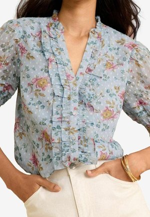 Femme portant un chemisier floral bleu clair avec des volants et des manches bouffantes, associé à un pantalon de couleur claire, mains dans les poches, et bracelets dorés au poignet.