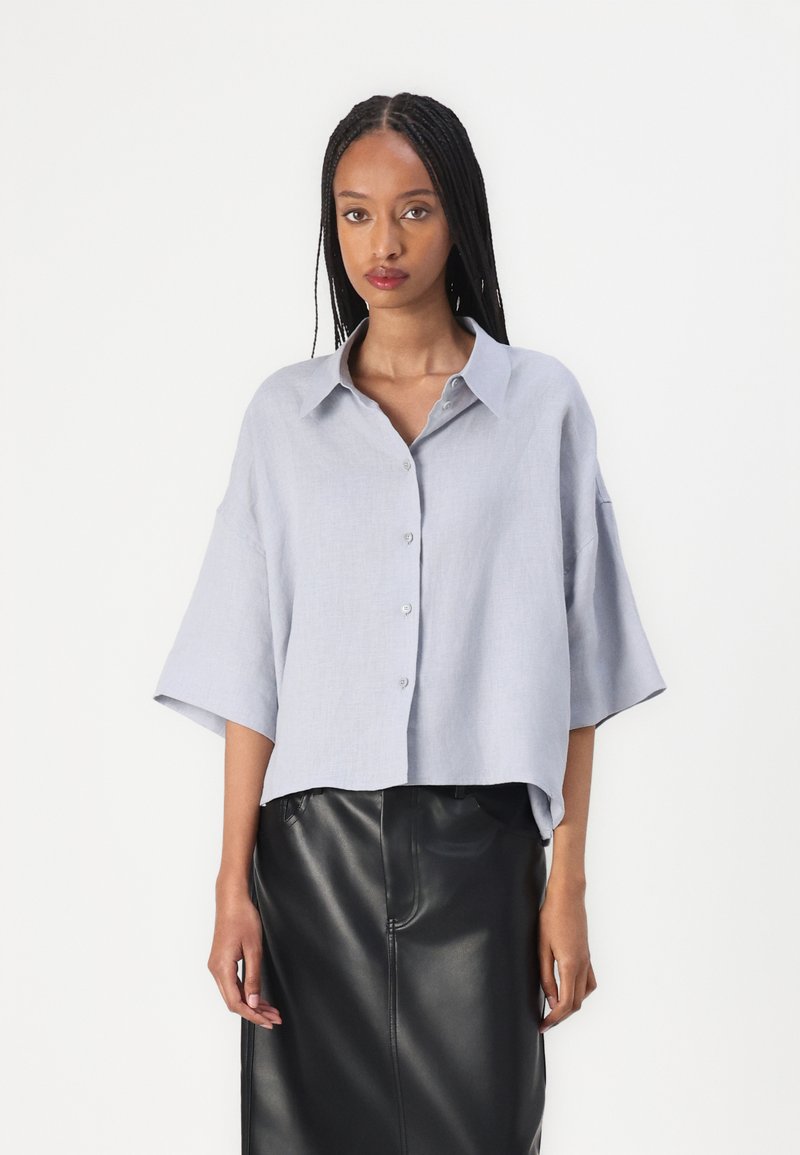 Camicia a maniche corte in lino azzurro chiaro con bottoni sul davanti, vestibilità ampia, colletto a punta e orlo dritto. Abbinata a una gonna di pelle nera.