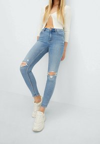 Jeans ajustados de tiro alto, de color azul claro, hechos de denim, con dos desgastes en las rodillas. Combinados con una chaqueta corta blanca y zapatillas beige.