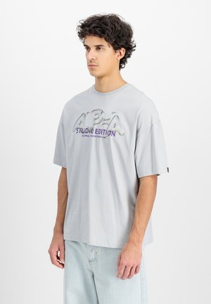 Svetlo siva oversize majica iz bombaža. Na prednji strani ima velik, sjajan, večbarvni logotip "ALPHA STUDIO EDITION". Kratki rukavi.