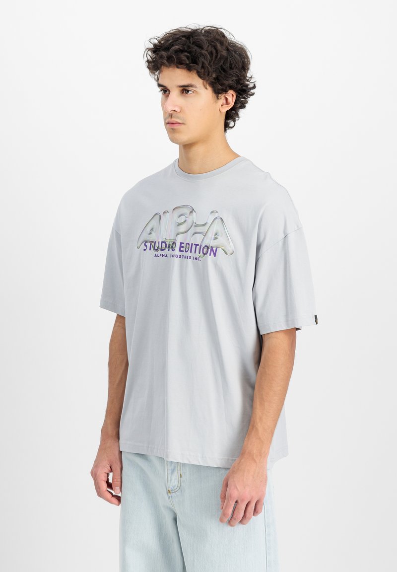 Tricou oversize gri deschis, fabricat din bumbac. Prezintă un logo mare, lucios și multicolor "ALPHA STUDIO EDITION" pe față. Mâneci scurte.