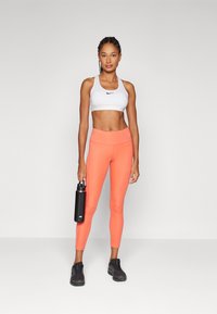 Bílý sportovní podprsenka s černým logem Nike, jasně oranžové legíny, černá láhev na vodu a černé sportovní boty. Hladký materiál, přiléhavý design.