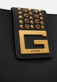 Musta tekonahkainen käsilaukku, jossa on kultainen "G"-logo ja koristeelliset kultaiset niitit sekä kirkkaat jalokivikoristeet läpän alueella.