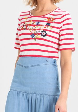 Femme portant une chemise à rayures rouges et blanches avec une broderie florale multicolore, associée à une jupe bleu clair taille haute.