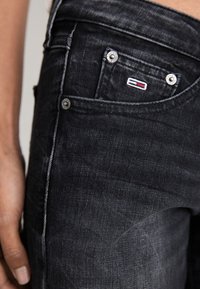 Jeans de mezclilla negra con un diseño de cintura media, dos bolsillos frontales, herrajes plateados y un pequeño logo en rojo, blanco y azul.