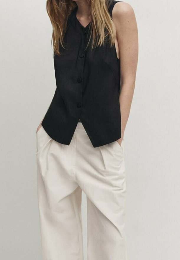 Femme portant un haut noir sans manches boutonné et un pantalon beige ample, les mains dans les poches, sur un fond uni.