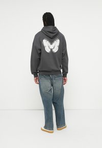 Felpa grigia con una grande stampa bianca di farfalla sulla schiena. Presenta un cappuccio con coulisse e una vestibilità comoda. Abbinata a jeans blu chiaro.