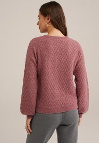 Roza, teksturirana pulover s herringbone vzorcem, okroglo izrezanino in balonastimi rokavi. Rebrasti manšeti in spodnji rob, v kombinaciji s sivimi hlačami.