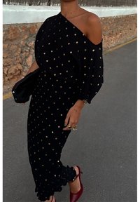 Femme portant une robe noire à une épaule ornée de points dorés, tenant une pochette noire et portant des chaussures rouges en marchant sur une rue pavée.