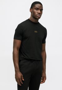 Μαύρο κοντομάνικο μπλουζάκι φτιαγμένο από βαμβάκι, με μια μικρή χρυσή κεντημένη λογότυπο «KARL LAGERFELD» στην αριστερή πλευρά του στήθους. Απλός, εφαρμοστός σχεδιασμός.