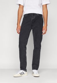 Črne jeans hlače s ravnim krojem, petimi žepi in diskretnim šivom. Nosi jih z belimi športnimi copati z sivimi detajli.