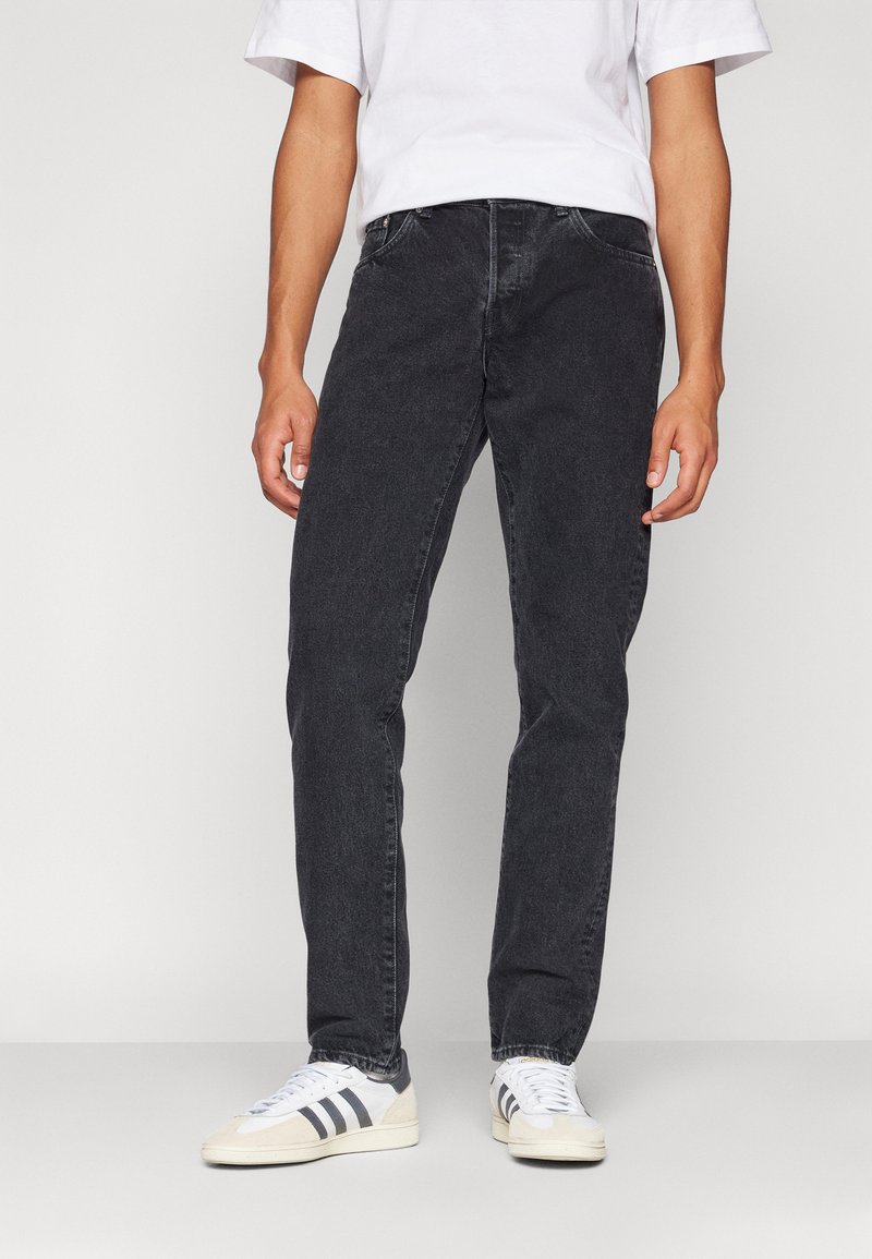 Črne jeans hlače s ravnim krojem, petimi žepi in diskretnim šivom. Nosi jih z belimi športnimi copati z sivimi detajli.