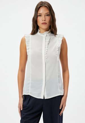 Blusa blanca sin mangas con acentos de volantes en los hombros y un cierre de botón, que presenta una tela suave y ligera y un cuello alto.