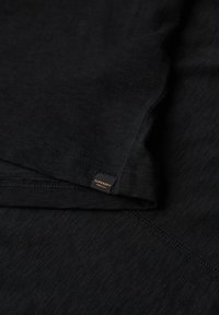 Nahaufnahme von schwarzem Stoff mit einem kleinen „Superdry Premium“-Label, das nahe dem Saum angenäht ist; sichtbare Nahtdetails.