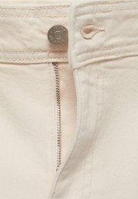 Gros plan sur un jean beige pâle ouvert avec un bouton métallique marqué MNG JEANS et une boutonnière cousue.