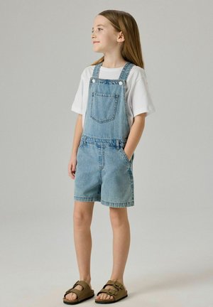 Ung jente står med hendene i lommene iført hvit T-skjorte, lyseblå denimshorts med seler, og brune sandaler med spenne på ensfarget bakgrunn.